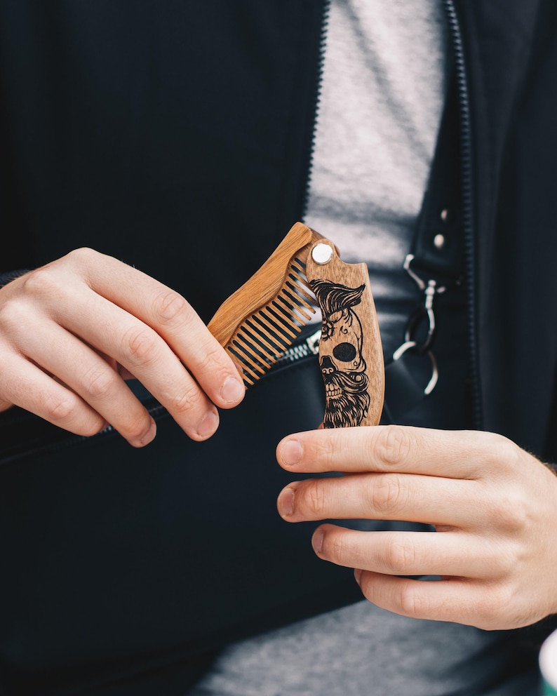 May include: A wooden folding beard comb with a skull design. The comb is made of light brown wood and features a detailed engraving of a skull with a beard and hair. The comb is held open, revealing the teeth of the comb.