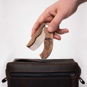 May include: A wooden folding comb with the words "REAL MAN" engraved on the handle. The comb is held open, revealing the teeth, above a brown leather toiletry bag. The comb is made of a light brown wood.