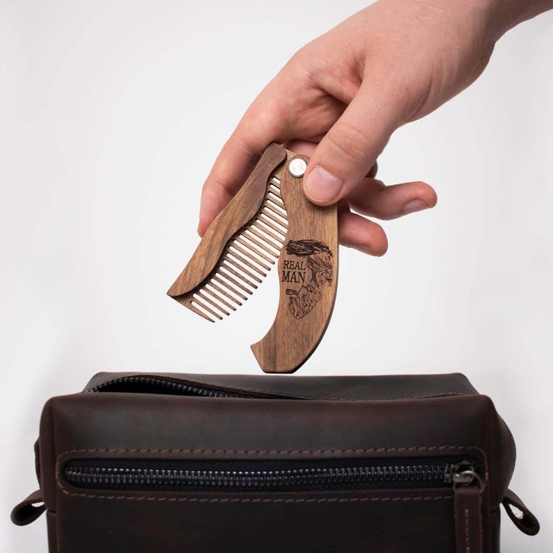 May include: A wooden folding comb with the words "REAL MAN" engraved on the handle. The comb is held open, revealing the teeth, above a brown leather toiletry bag. The comb is made of a light brown wood.