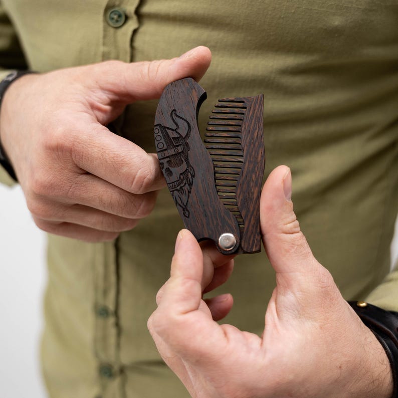 May include: A dark wood folding beard comb with a Viking skull design. The comb is held open, revealing the teeth. The handle features a detailed engraving of a Viking skull wearing a horned helmet and a long beard.