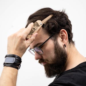May include: A wooden folding comb with a detailed engraved design of a bearded face. The comb is being used on dark brown hair. The comb is light brown and has a magnetic closure. The person in the image is wearing glasses and a black shirt.