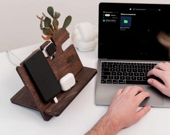 May include: A dark brown wooden desk organizer holds a black smartphone, a silver smartwatch, and white earbuds. The organizer has a slanted design and the name "SMITH" is engraved on it. A laptop is open in the background.