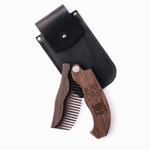 May include: A dark brown wooden folding comb, engraved with the words "SUPER DAD" and a superhero mask silhouette. It is presented with a black leather case for storage and protection.