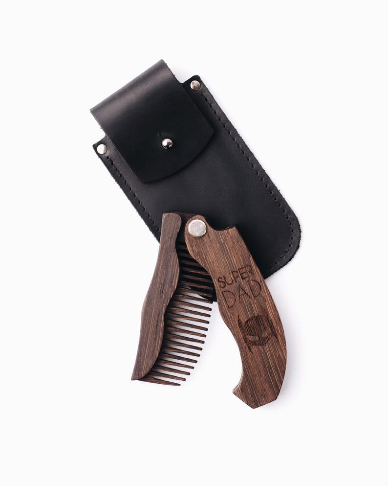 May include: A dark brown wooden folding comb, engraved with the words "SUPER DAD" and a superhero mask silhouette. It is presented with a black leather case for storage and protection.