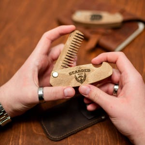 May include: A wooden pocket comb with a bottle opener and the text "The Bearded Man" engraved on it. The comb is open and being held by a person's hands.