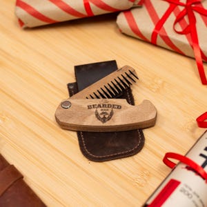 May include: A wooden beard comb with a leather case. The comb has a metal hinge and a small metal circle on the side. The comb is engraved with the words "The Bearded Man".