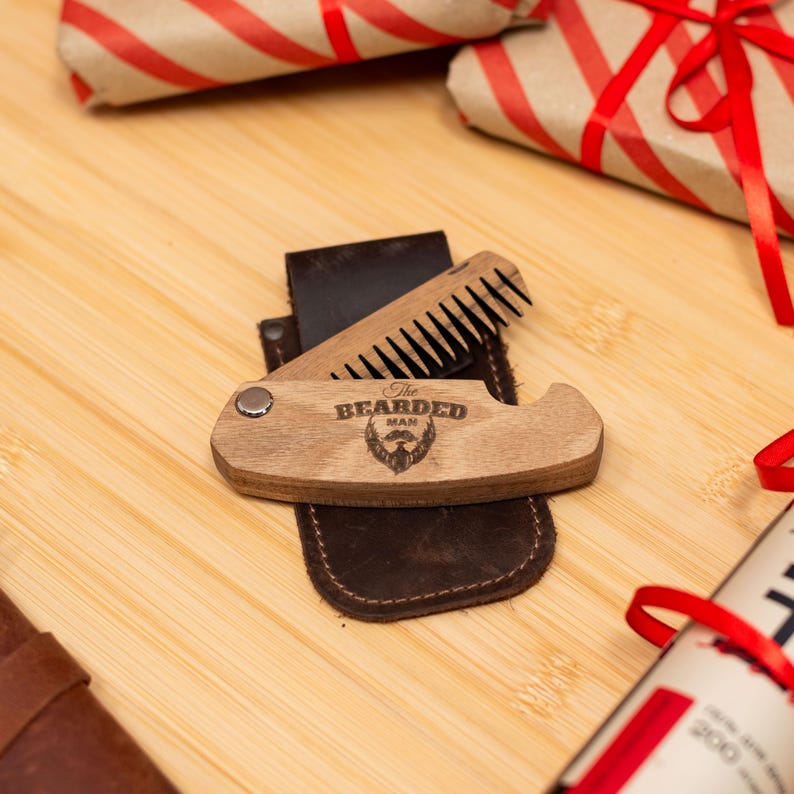 May include: A wooden beard comb with a leather case. The comb has a metal hinge and a small metal circle on the side. The comb is engraved with the words "The Bearded Man".