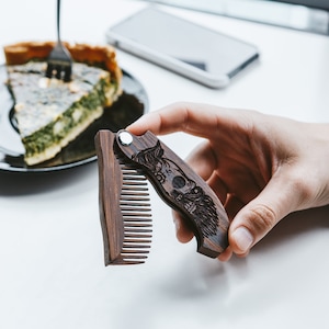 May include: A folding wooden comb with a carved skull and beard design. The brown comb is open, showing the teeth. It has a silver hinge. The comb is held in a hand. The comb is a grooming accessory.