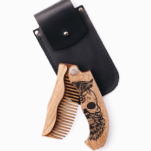 May include: A wooden folding beard comb with a skull design and a black leather case. The comb is made of light brown wood and features a detailed engraved skull with a mustache and beard. The black case has a snap closure.