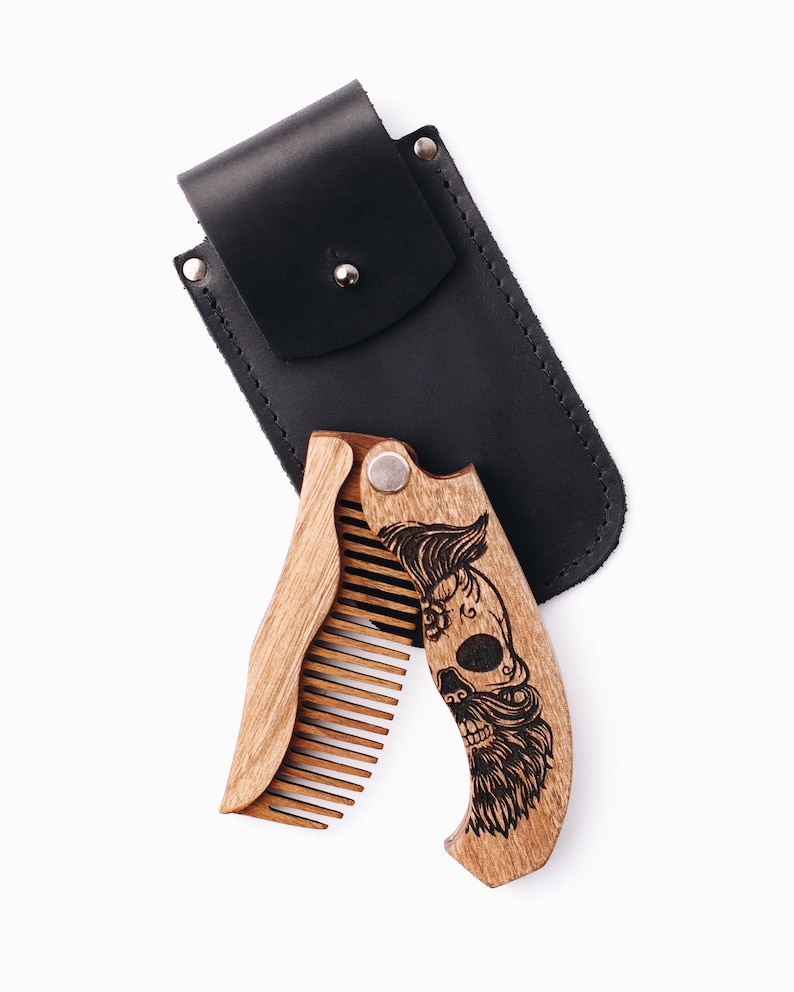 May include: A wooden folding beard comb with a skull design and a black leather case. The comb is made of light brown wood and features a detailed engraved skull with a mustache and beard. The black case has a snap closure.