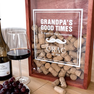 May include: A rectangular wooden shadow box with a glass front filled with natural corks. The glass features white text reading "GRANDPA'S GOOD TIMES A toast to the memories" with a mustache graphic and "GRAG est. 2026" below. A wine glass and grapes are nearby.