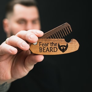 Könnte beinhalten: Ein Holzbartkamm mit einem Flaschenöffner. Der Kamm ist geöffnet und der Flaschenöffner ist sichtbar. Der Kamm trägt den Text "Fear the Beard" und ein Bart-Symbol ist eingraviert.