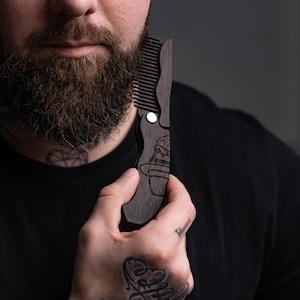 May include: A dark brown wooden beard comb with a detailed carved design. The comb is folded and held in the hand, showcasing the intricate craftsmanship. The comb is designed for grooming and styling facial hair.