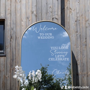 May include: An arched floor mirror with a gold frame featuring white text that reads Welcome TO OUR WEDDING, YOU LOOK Stunning LET'S CELEBRATE, Charly & Tony, and 25.5.24. Two clear glass vases with white orchid plants sit in front of the mirror.