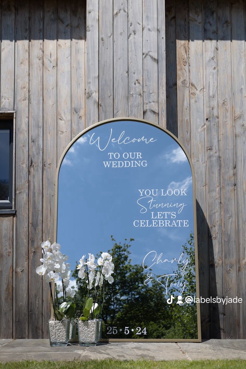 May include: An arched floor mirror with a gold frame featuring white text that reads Welcome TO OUR WEDDING, YOU LOOK Stunning LET'S CELEBRATE, Charly & Tony, and 25.5.24. Two clear glass vases with white orchid plants sit in front of the mirror.