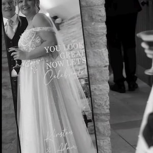May include: An arched floor mirror featuring white vinyl lettering that reads Welcome TO OUR WEDDING, YOU LOOK GREAT NOW LETS Celebrate, Kirsten & Adam, and 14.02.2025. The mirror reflects a person in a wedding dress and a person in a suit.