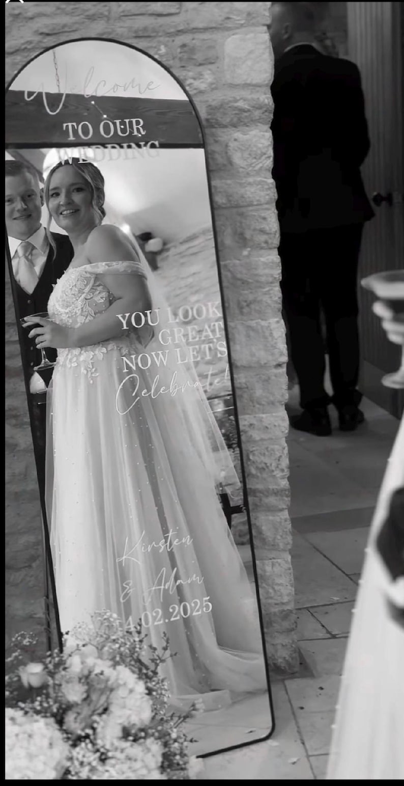 May include: An arched floor mirror featuring white vinyl lettering that reads Welcome TO OUR WEDDING, YOU LOOK GREAT NOW LETS Celebrate, Kirsten & Adam, and 14.02.2025. The mirror reflects a person in a wedding dress and a person in a suit.