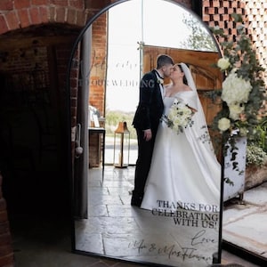May include: An arched floor mirror with a black metal frame featuring white text that reads "WELCOME TO OUR WEDDING" at the top and "THANKS FOR CELEBRATING WITH US! Love Mr & Mrs Flower" at the bottom, reflecting a couple kissing in a brick building.