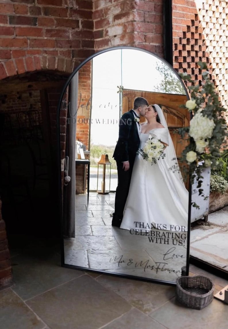 May include: An arched floor mirror with a black metal frame featuring white text that reads "WELCOME TO OUR WEDDING" at the top and "THANKS FOR CELEBRATING WITH US! Love Mr & Mrs Flower" at the bottom, reflecting a couple kissing in a brick building.