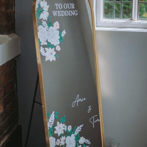 May include: An arched floor mirror with a gold frame featuring white floral illustrations and the text Welcome TO OUR WEDDING and the names Amie & Tom in white script. The mirror is displayed on a stand, showcasing decorative white flowers.