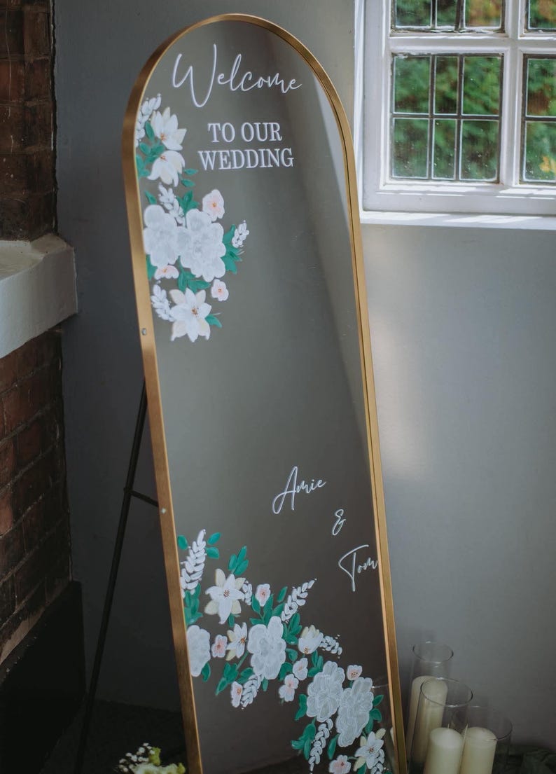 May include: An arched floor mirror with a gold frame featuring white floral illustrations and the text Welcome TO OUR WEDDING and the names Amie & Tom in white script. The mirror is displayed on a stand, showcasing decorative white flowers.