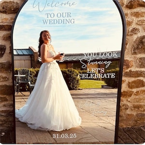 May include: An arched floor mirror with a black frame featuring white text that reads Welcome TO OUR WEDDING, YOU LOOK Stunning LET'S CELEBRATE, Vicki & Tommy, and 31.03.25. The mirror reflects a person in a white wedding dress holding a glass.