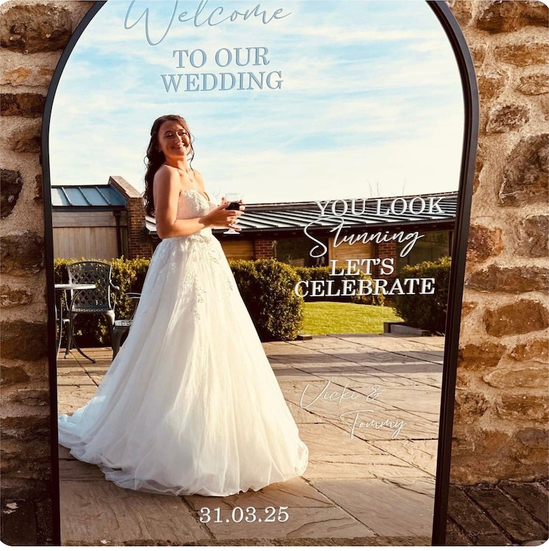 May include: An arched floor mirror with a black frame featuring white text that reads Welcome TO OUR WEDDING, YOU LOOK Stunning LET'S CELEBRATE, Vicki & Tommy, and 31.03.25. The mirror reflects a person in a white wedding dress holding a glass.