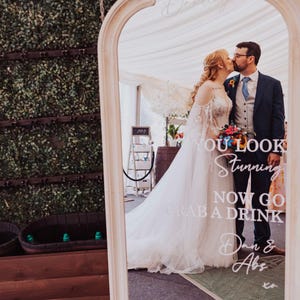 May include: A tall, arched floor mirror with a white frame featuring white vinyl lettering that reads Hello Darling, YOU LOOK Stunning NOW GO GRAB A DRINK, Dan & Abs xo, and 12.04.2025. The mirror reflects a couple kissing in formal wedding attire.