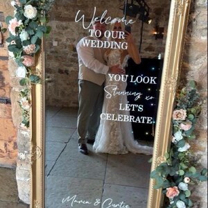 May include: A large rectangular mirror with an ornate gold frame, decorated with artificial eucalyptus and pink and white flowers. The glass features white text reading Welcome TO OUR WEDDING, YOU LOOK Stunning xo LET'S CELEBRATE, Maria & Curtis, 15.07.2026.
