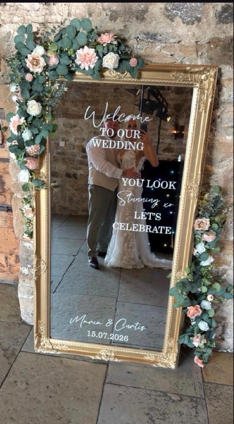 May include: A large rectangular mirror with an ornate gold frame, decorated with artificial eucalyptus and pink and white flowers. The glass features white text reading Welcome TO OUR WEDDING, YOU LOOK Stunning xo LET'S CELEBRATE, Maria & Curtis, 15.07.2026.