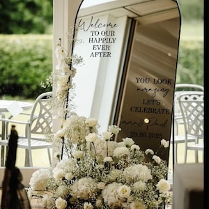 May include: An arched floor mirror with a black frame featuring black vinyl text that reads Welcome TO OUR HAPPILY EVER AFTER, YOU LOOK amazing LET'S CELEBRATE, Emma & Paul, and 29.05.2025. A large arrangement of white flowers and baby's breath sits at the base.