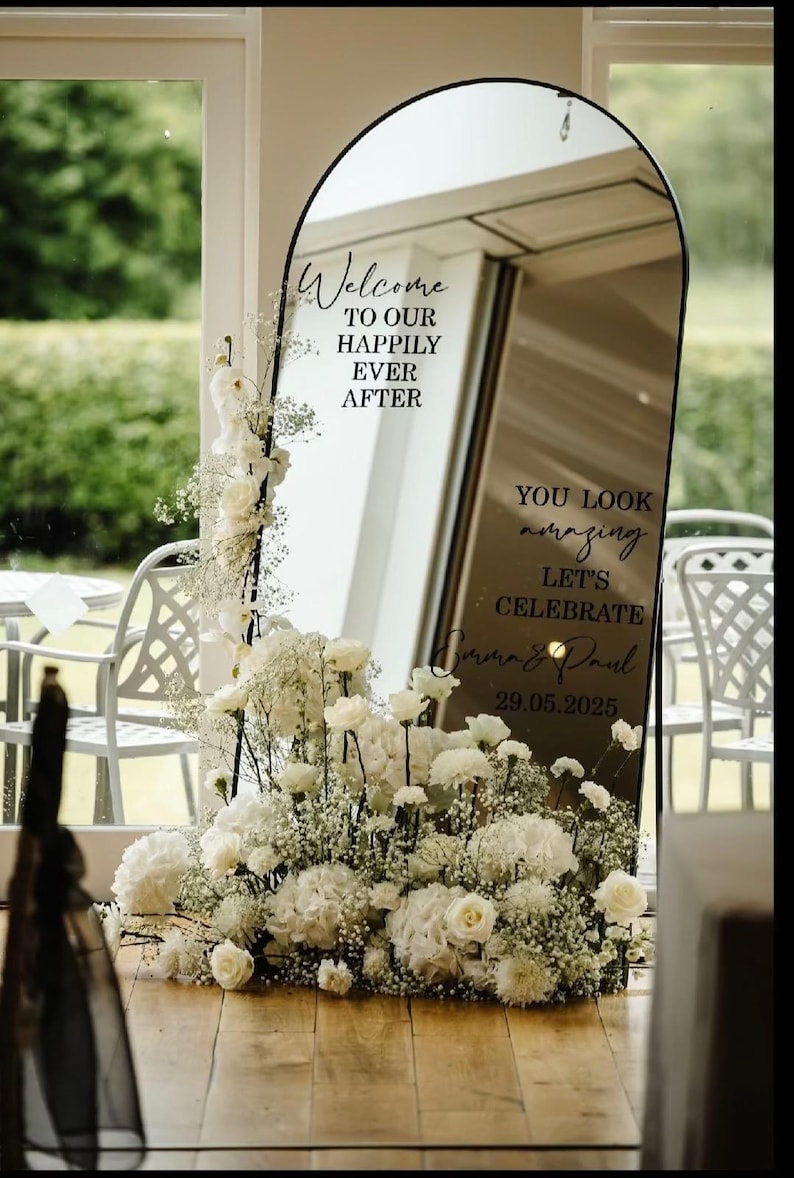 May include: An arched floor mirror with a black frame featuring black vinyl text that reads Welcome TO OUR HAPPILY EVER AFTER, YOU LOOK amazing LET'S CELEBRATE, Emma & Paul, and 29.05.2025. A large arrangement of white flowers and baby's breath sits at the base.
