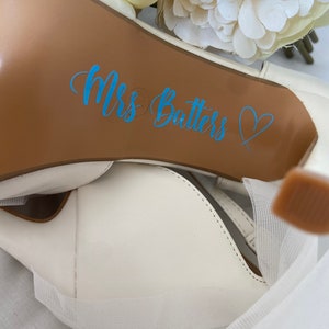 May include: White wedding shoe with a brown sole. The sole has a blue sticker that says "Mrs. Batters" with a heart.
