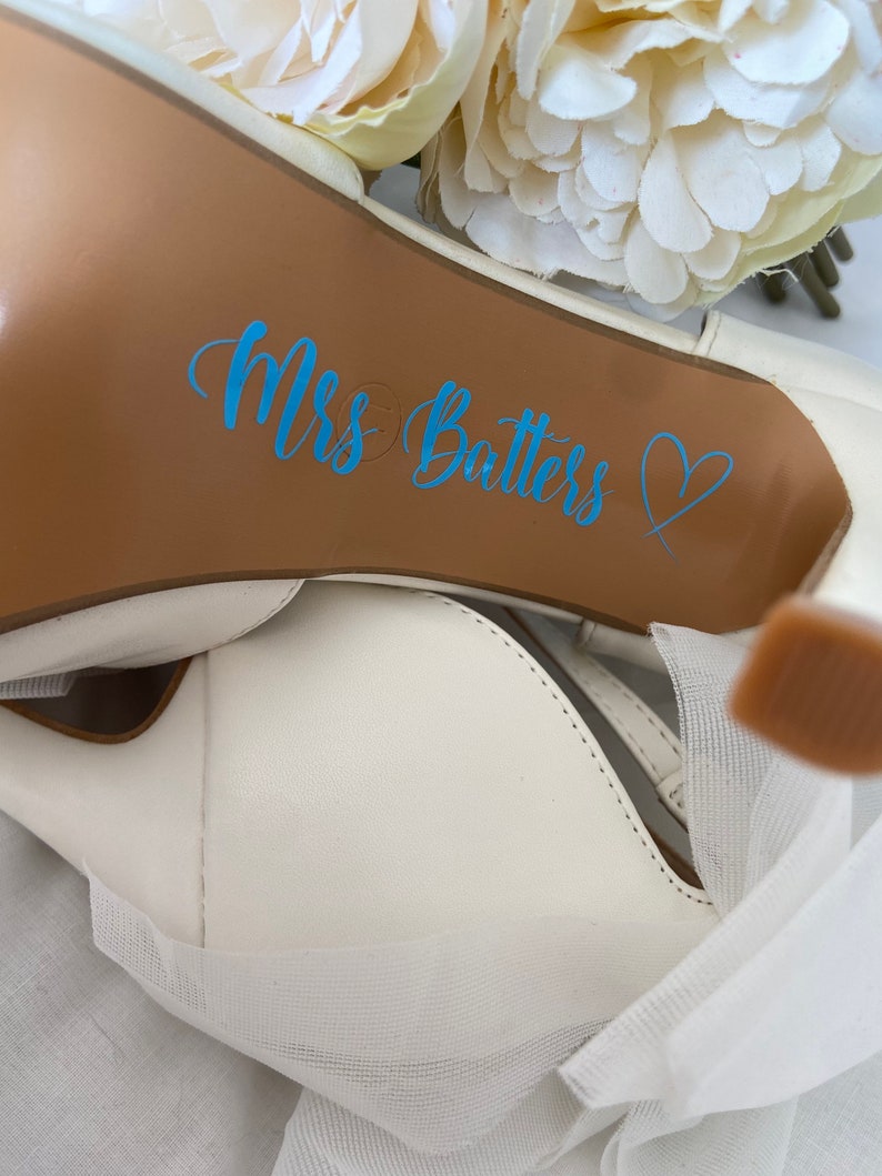May include: White wedding shoe with a brown sole. The sole has a blue sticker that says "Mrs. Batters" with a heart.