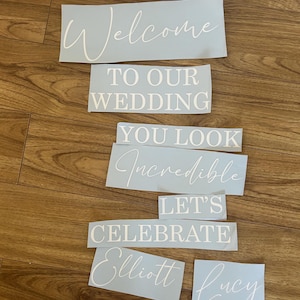 May include: A set of light blue vinyl decals featuring white text that reads: Welcome TO OUR WEDDING YOU LOOK Incredible LET'S CELEBRATE Elliott Lucy 02/09/2025. The lettering uses a combination of elegant script and bold block fonts.