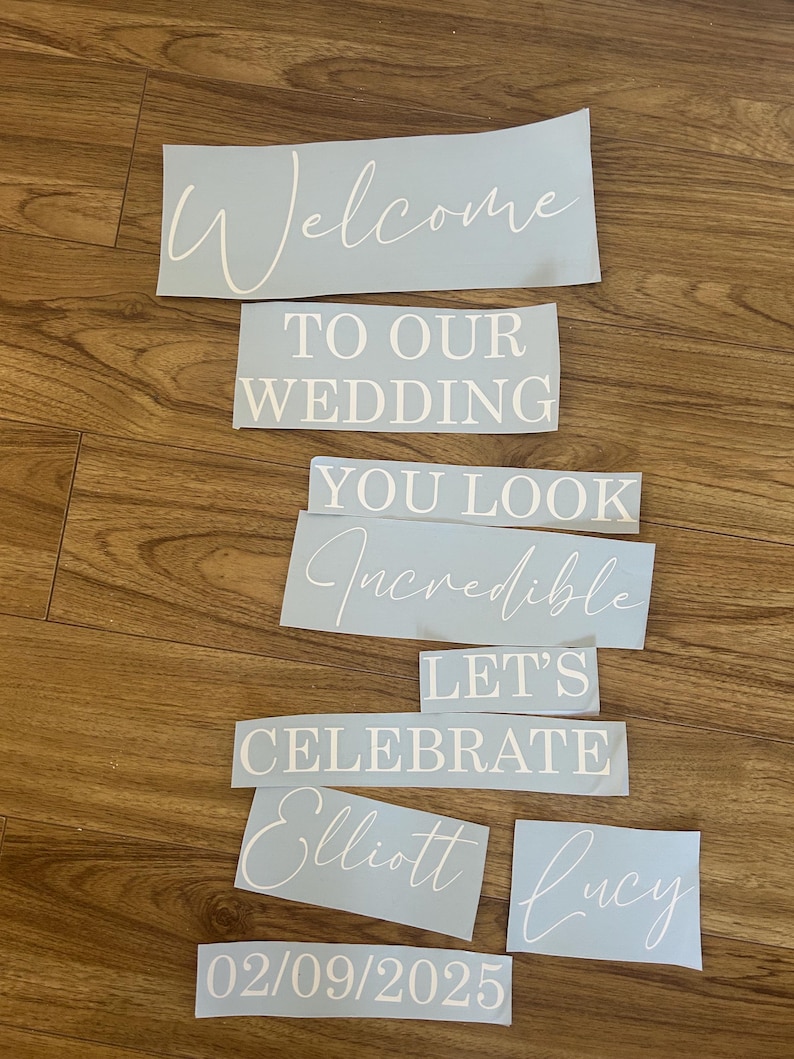 May include: A set of light blue vinyl decals featuring white text that reads: Welcome TO OUR WEDDING YOU LOOK Incredible LET'S CELEBRATE Elliott Lucy 02/09/2025. The lettering uses a combination of elegant script and bold block fonts.