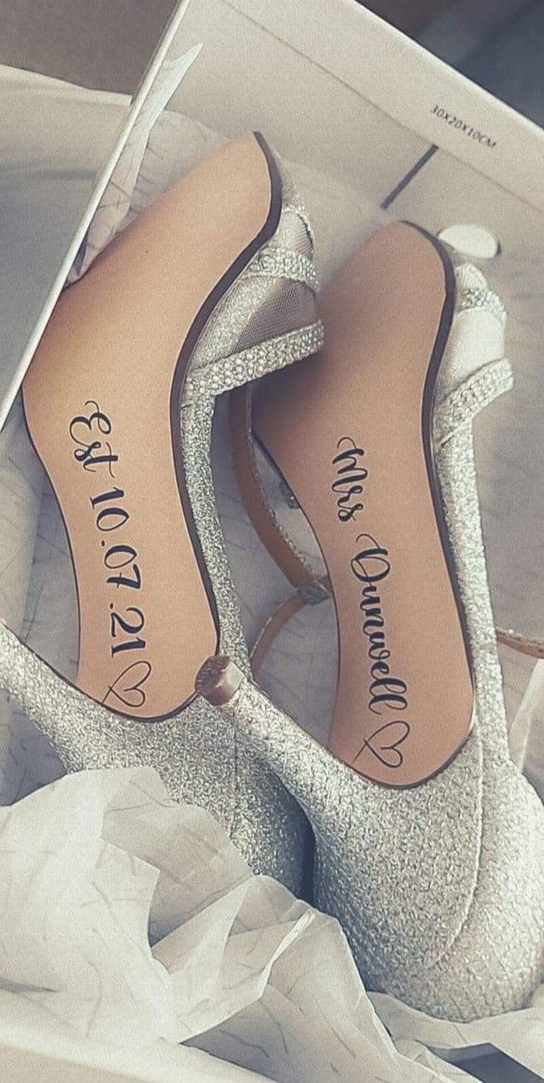 May include: A pair of silver glitter high heel shoes with the text "Mrs. Dunwell" and "Est. 10.07.21" written on the soles.