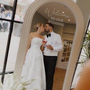 May include: An arched floor mirror with a light-colored frame featuring the white text Welcome TO OUR WEDDING. The mirror reflects a person in a strapless white gown and a person in a white shirt, black bow tie, and black trousers kissing.