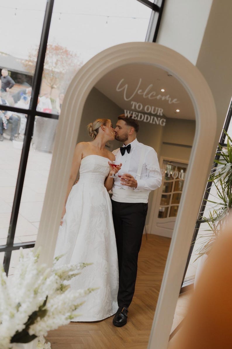 May include: An arched floor mirror with a light-colored frame featuring the white text Welcome TO OUR WEDDING. The mirror reflects a person in a strapless white gown and a person in a white shirt, black bow tie, and black trousers kissing.