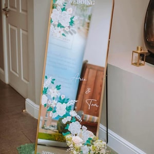 May include: An arched floor mirror with a gold frame featuring white floral decals and white text that reads Welcome TO OUR WEDDING, Amie & Tom, and 30TH JULY. The mirror is decorated with white and pink flowers in glass vases at the base.