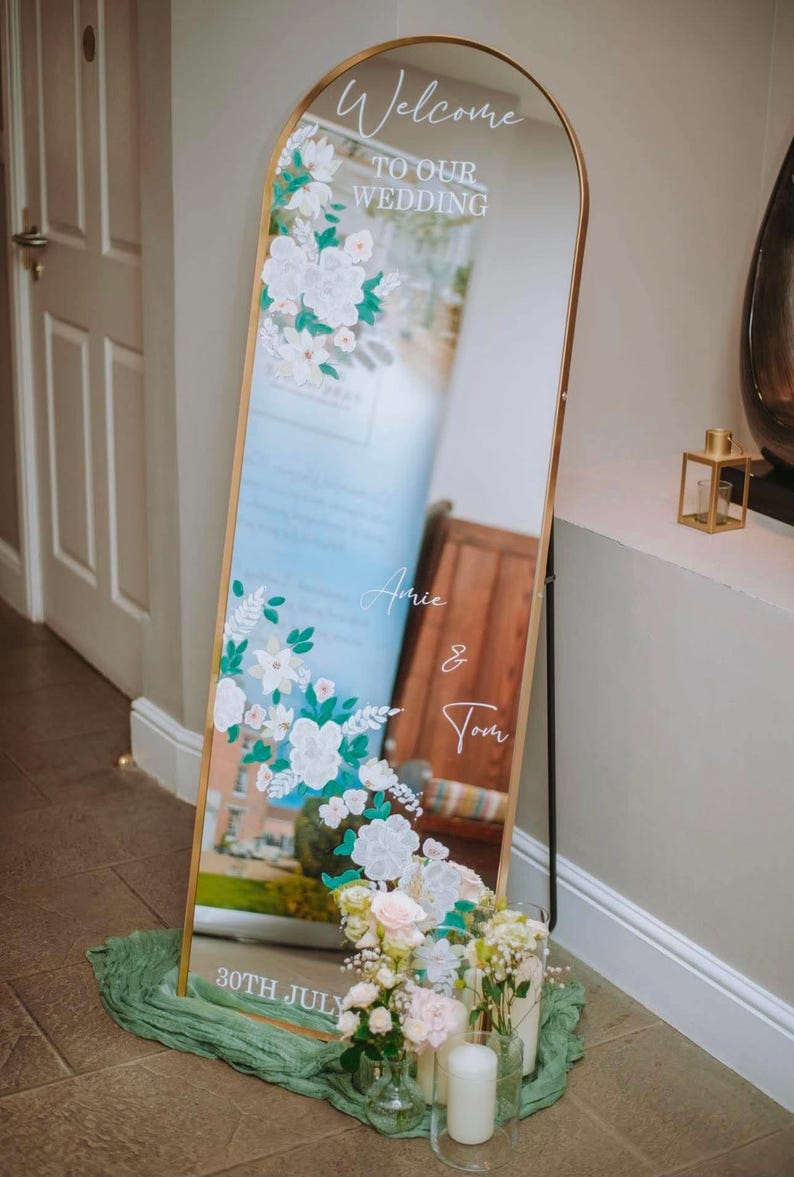 May include: An arched floor mirror with a gold frame featuring white floral decals and white text that reads Welcome TO OUR WEDDING, Amie & Tom, and 30TH JULY. The mirror is decorated with white and pink flowers in glass vases at the base.