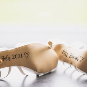 May include: A pair of white bridal shoes with the date "31st July 2021" and a heart written on the sole of one shoe and the text "Mrs. Summer" and a heart written on the sole of the other shoe.