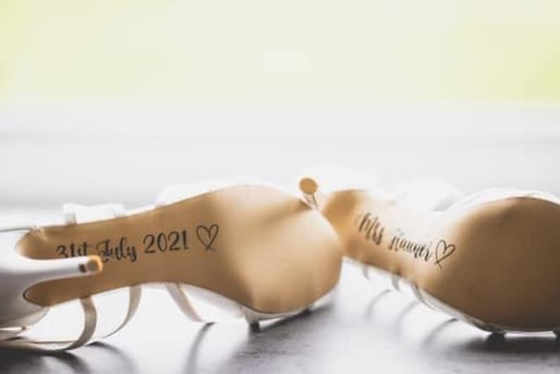 May include: A pair of white bridal shoes with the date "31st July 2021" and a heart written on the sole of one shoe and the text "Mrs. Summer" and a heart written on the sole of the other shoe.