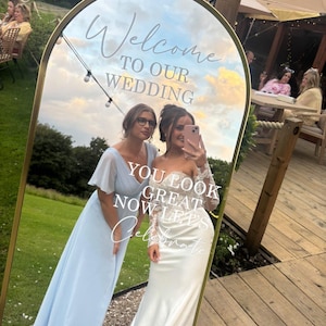 May include: An arched floor mirror with a gold frame featuring white text that reads Welcome TO OUR WEDDING and YOU LOOK GREAT NOW LETS Celebrate. The mirror reflects an outdoor scene with a grassy lawn, wooden decking, and string lights.