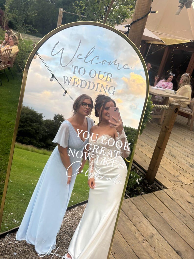 May include: An arched floor mirror with a gold frame featuring white text that reads Welcome TO OUR WEDDING and YOU LOOK GREAT NOW LETS Celebrate. The mirror reflects an outdoor scene with a grassy lawn, wooden decking, and string lights.