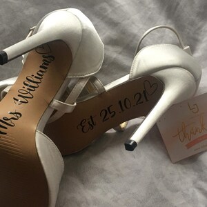 May include: A pair of white high-heeled shoes with the text "Mrs Williams" and "Est 25.10.21" written on the soles. A small white card with the text "thank you" is visible in the background.