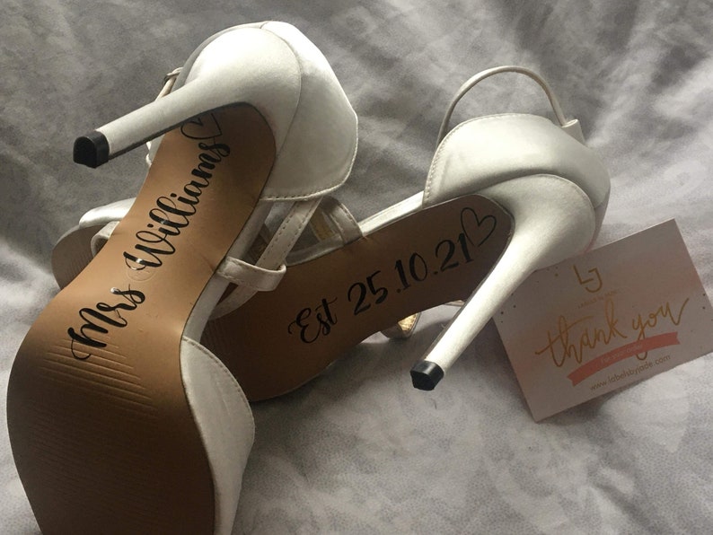May include: A pair of white high-heeled shoes with the text "Mrs Williams" and "Est 25.10.21" written on the soles. A small white card with the text "thank you" is visible in the background.