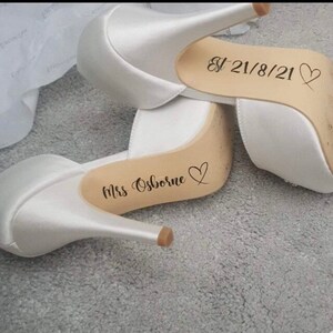 May include: A pair of white satin high heel shoes with personalized messages on the soles. One shoe says "Mrs. Osborne" with a heart and the other says "Est 21/8/21" with a heart.