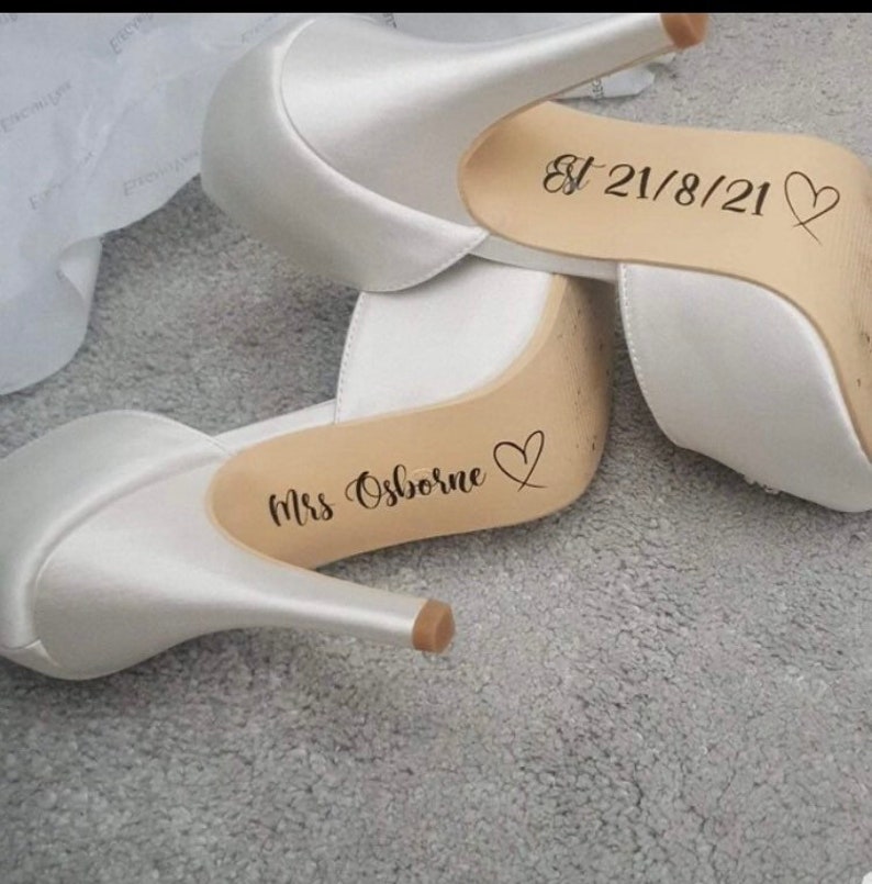May include: A pair of white satin high heel shoes with personalized messages on the soles. One shoe says "Mrs. Osborne" with a heart and the other says "Est 21/8/21" with a heart.