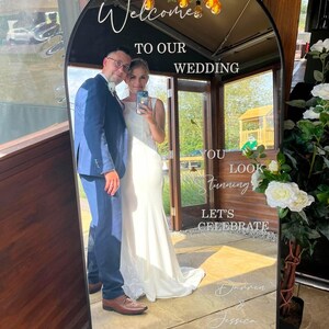 May include: An arched floor mirror with a black frame featuring white text that reads Welcome TO OUR WEDDING YOU LOOK Stunning LET'S CELEBRATE Darren & Jessica 27.06.2026. The mirror reflects a person in a blue suit and a person in a white dress.